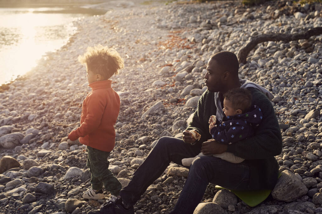 Pappa sitter på strand med bebis i knät och son bredvid