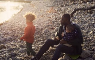 Pappa sitter på strand med bebis i knät och son bredvid